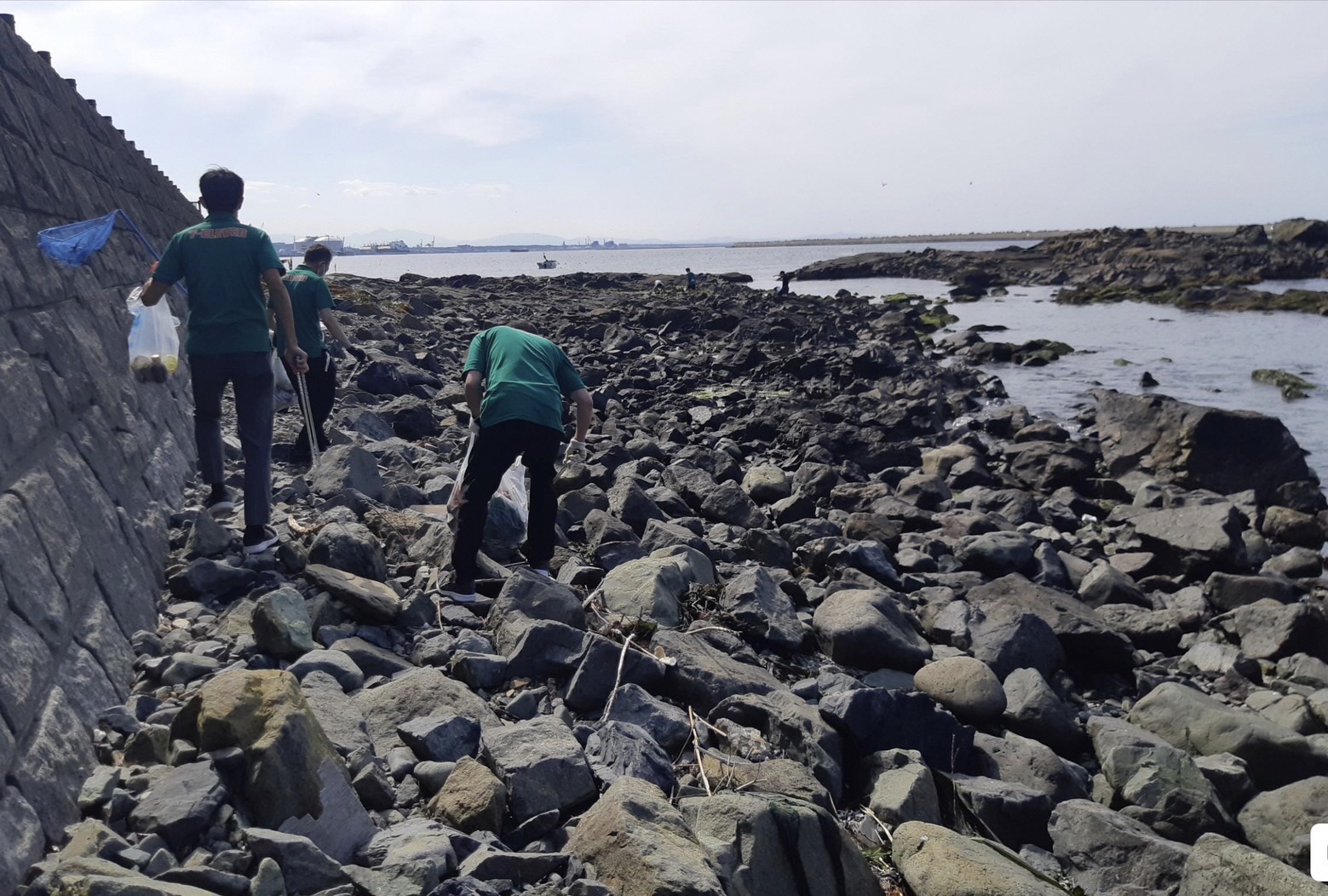 海と道路の間の岩場に降りて、岩の間のごみを拾っている三人の写真