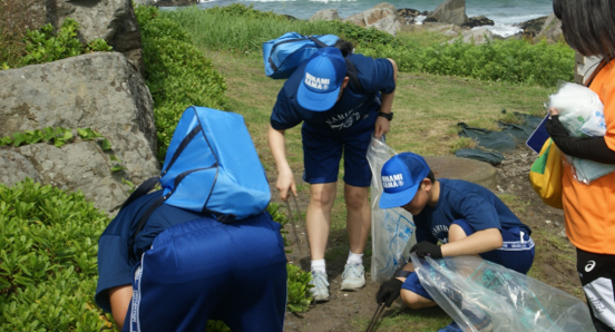 海を背景に草の生えている場所のごみを拾っている生徒三人と職員の写真