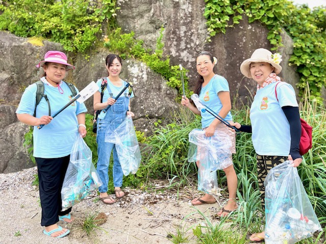 大きな岩場の前で、トングでごみをつかんでいる4人の女性の写真。手にはゴミの入ったボランティア用ごみ袋。