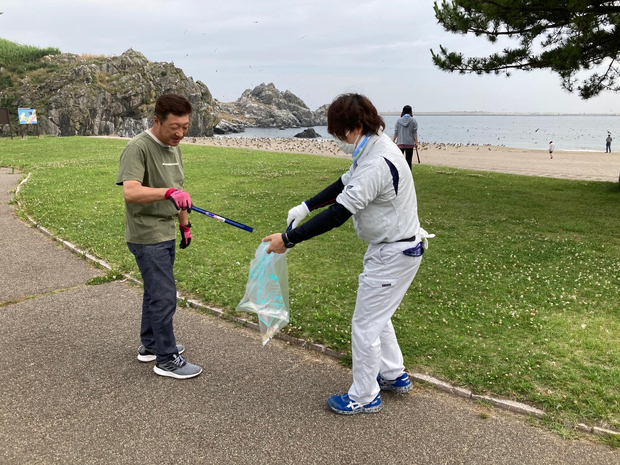 うみねこと海の見える場所。トングでごみをつかんでいる男性とごみ袋を広げている男性の写真。