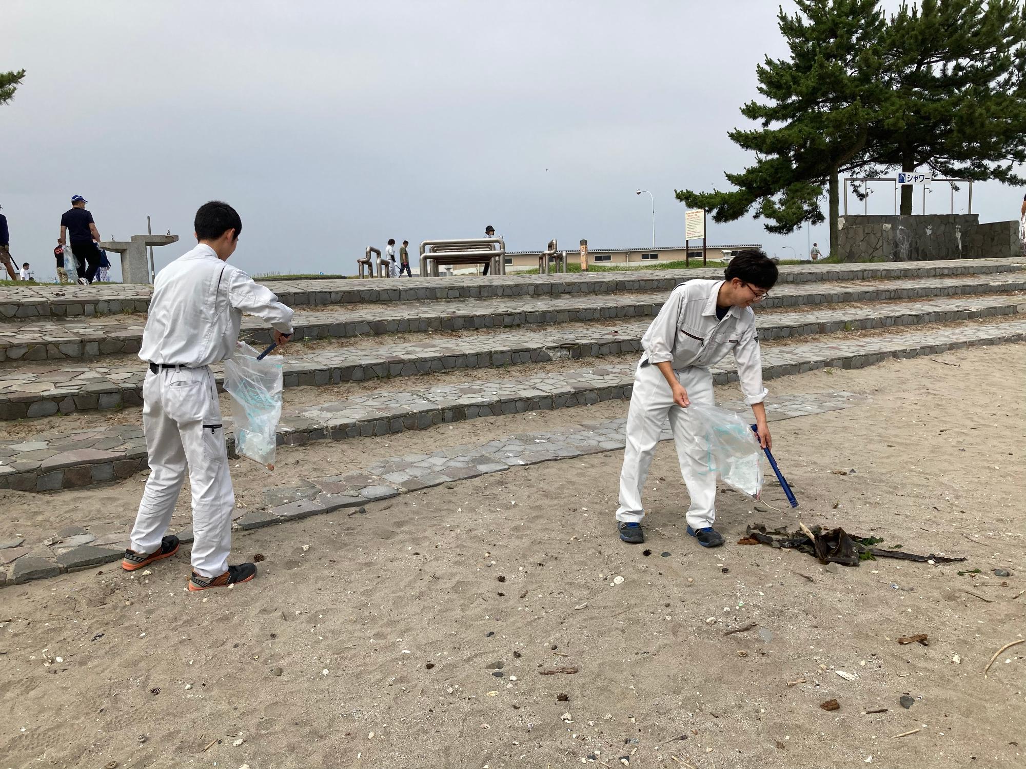 蕪島海岸の砂浜にある階段。トングで砂の中のごみを拾っている男性2人の写真。