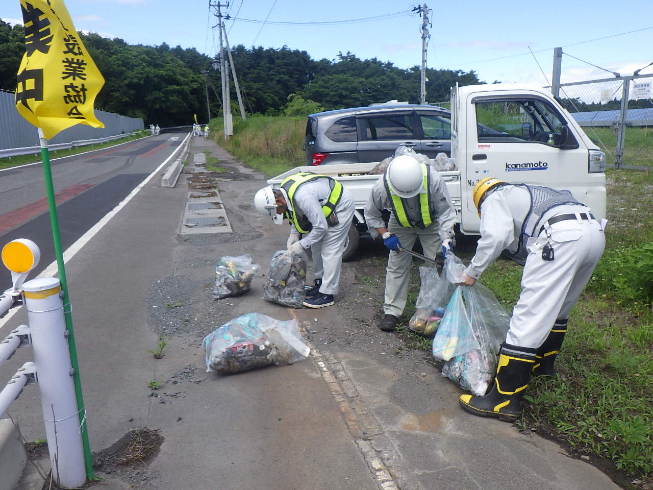 作業服に反射テープのついたベストを身に着け、道路脇のごみを袋に詰めている男性3人の写真