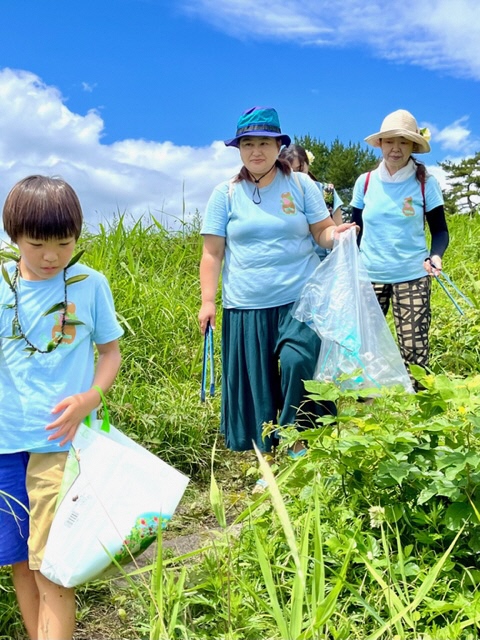 女性二人、男児1人。膝位の丈の草の生えている場所をボランティア用ごみ袋を手に持って一列に並んでいる写真