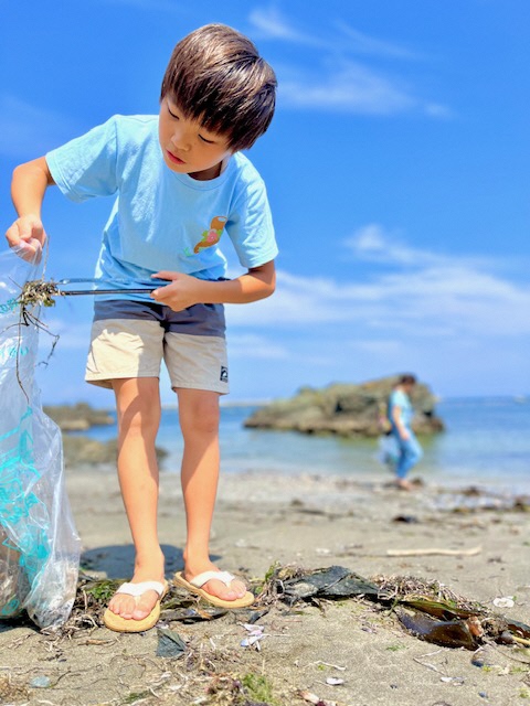 青い空と海をバックに、トングでゴミをつかんでいる男児の写真