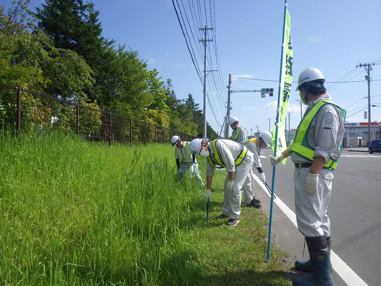 道路脇の草の生えている場所をのぼりをたてて、トングでごみを拾っている写真