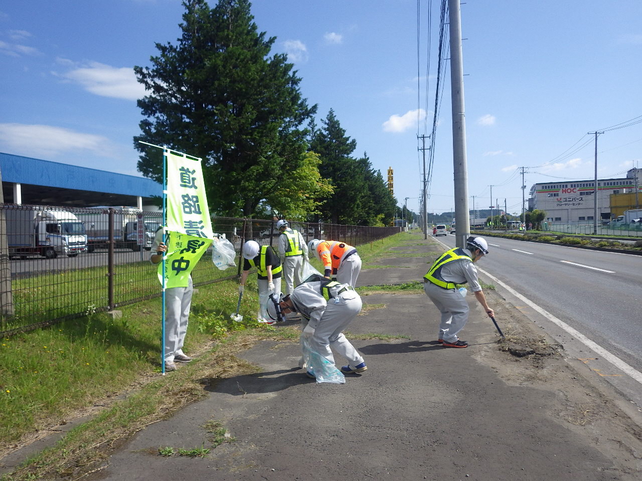 のぼりをたて、作業服に反射ベストを着用し歩道のごみを数名で拾っている写真