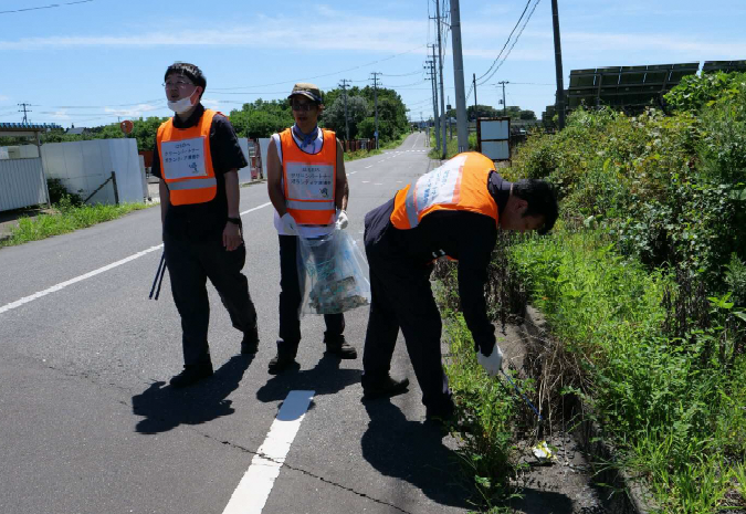 オレンジ色のビブスを着用し、道路のごみを拾っている男性三人の写真