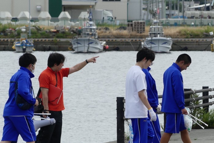 漁船の見える場所で活動する、トレーニングウエアの生徒4人と職員の男性の写真