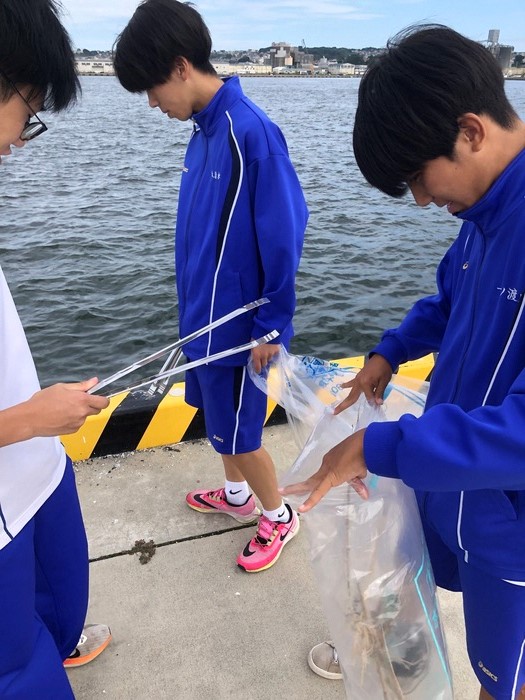 漁港の海上ぎりぎりの場所でトングを持つ生徒、ボランティア用ごみ袋を持つ生徒の写真