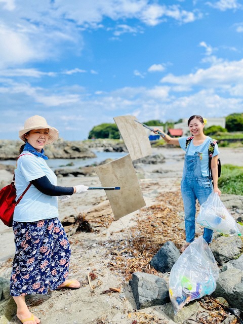 青空の広がる砂浜で拾ったごみをトングで挟み、こちらに見せている女性二人の写真