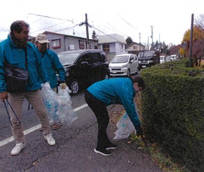 博物館前の植え込みの下のごみをトングで拾っている人、その後ろにいる二人の写真