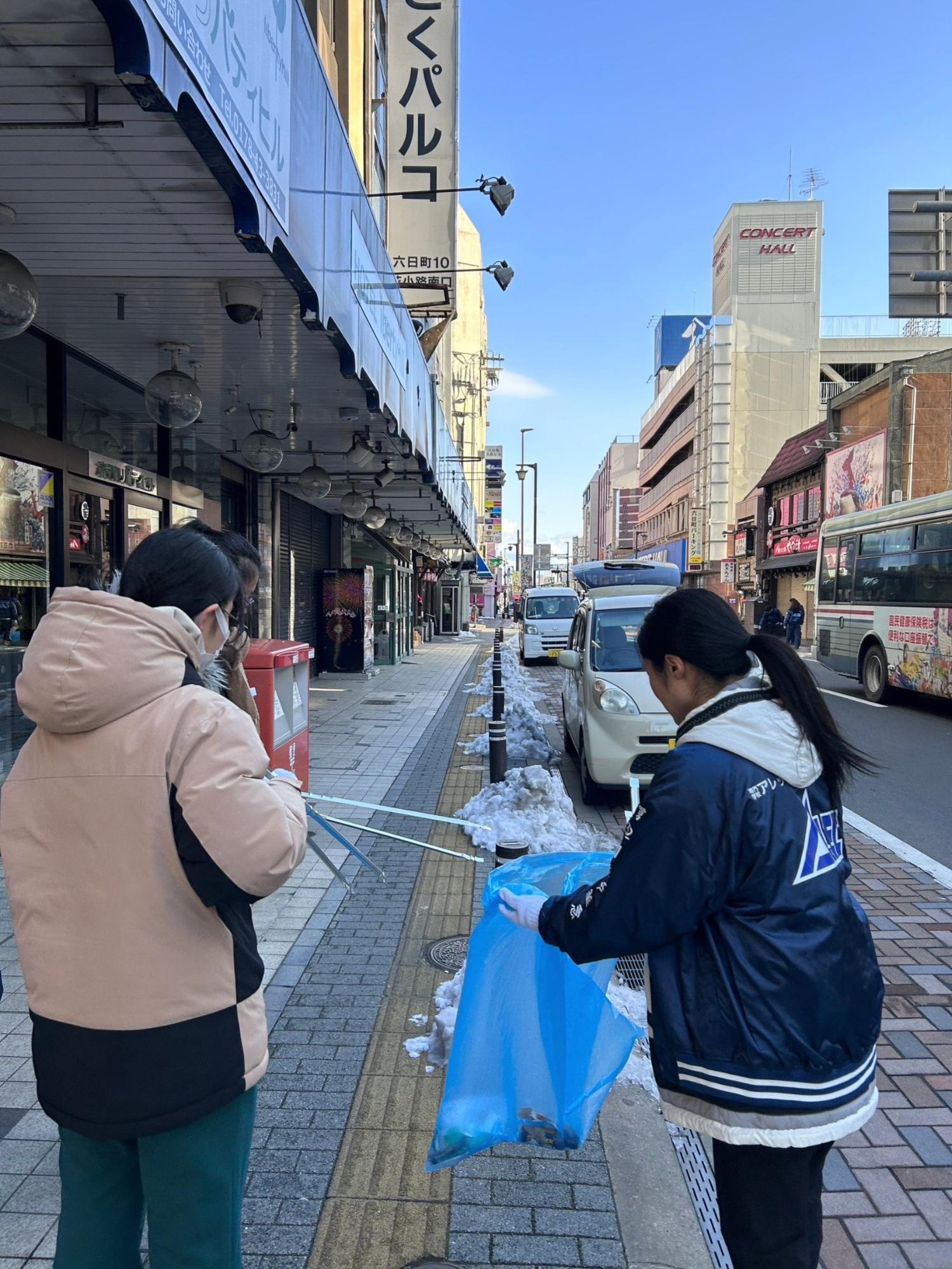 六日町の歩道のごみを女性二人でトングで掴んでごみ袋に入れている写真。