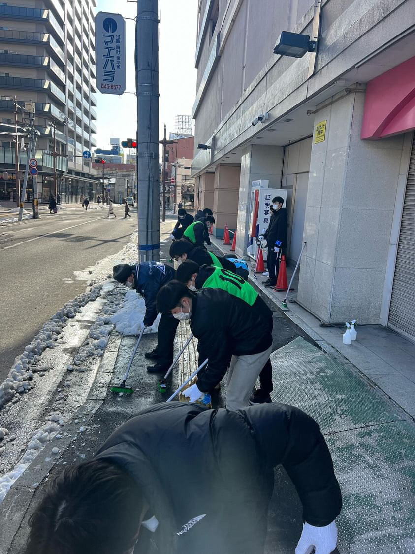 さくら野百貨店の脇の歩道をデッキブラシで擦り清掃している、学生数名の写真。道路には雪が見えている。