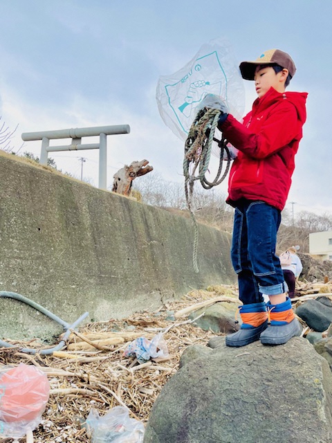 鳥居の見える堤防下で漂着したひも状のごみを袋に入れている男児の写真