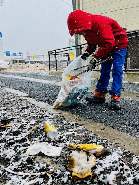防寒着のフードをかぶり、雪化粧した歩道に落ちている軍手や空き缶をトングで拾う男児の写真