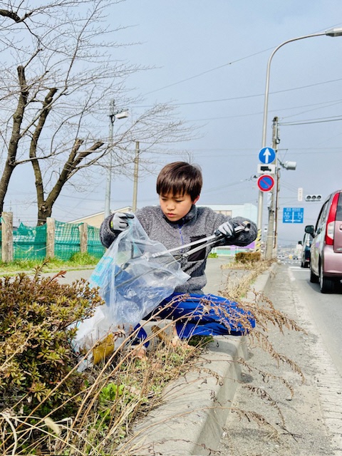車道のすぐ脇の歩道で、トングとボランティア用ごみ袋を持ち拾ったごみを入れている男児の写真