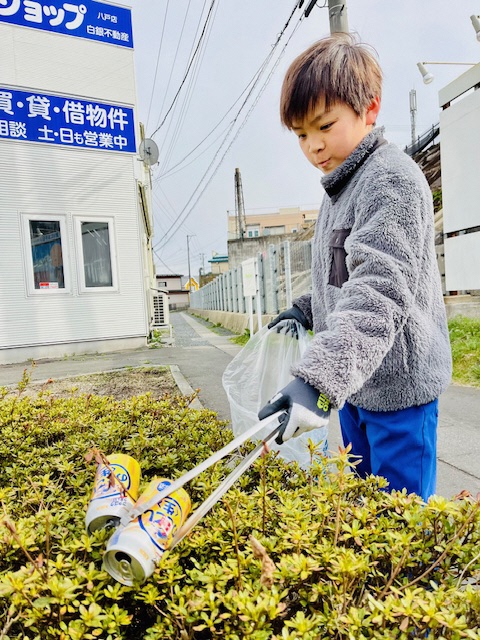 歩道の植え込みの上にポイ捨てされた酒類缶をトングではさみ拾っている男児の写真
