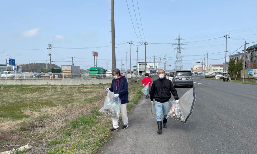 青空の中道路脇のごみを集めている写真。手に持ったボランティア用ごみ袋には空き缶がたくさん入っている。