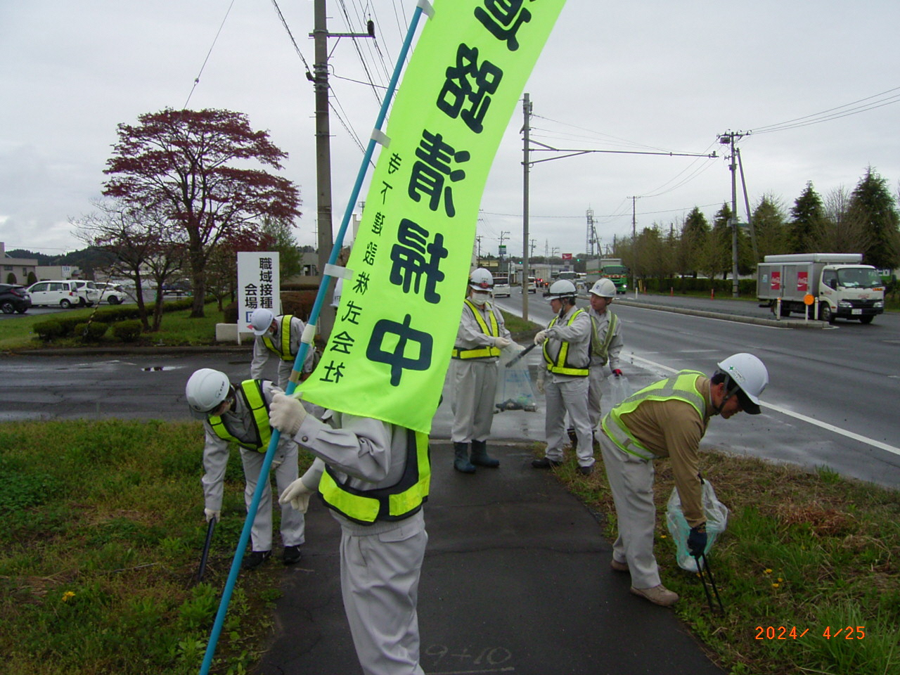 のぼり旗を掲げて、作業服に光るビブスを着用しごみを拾う人たちの写真