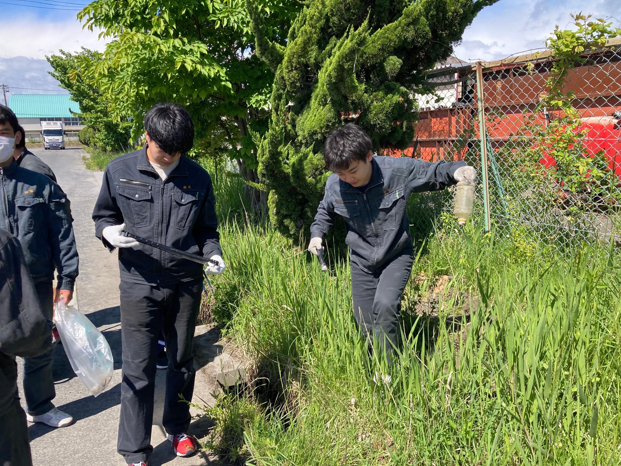 フェンス下の雑草の生い茂る場所のごみを拾っている数名の写真