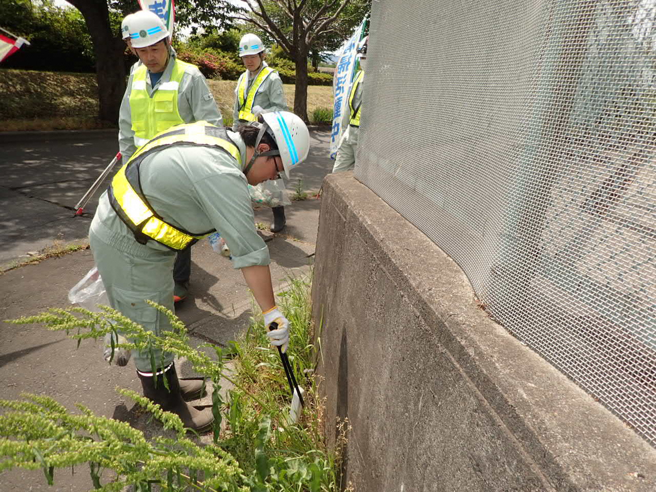 歩道の雑草の中からトングでごみを拾っている作業着の人の写真