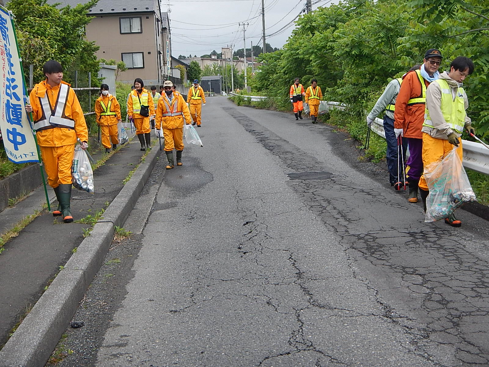 オレンジ色の作業服に清掃活動中の幟旗を掲げ、住宅地の歩道のごみを集めている写真