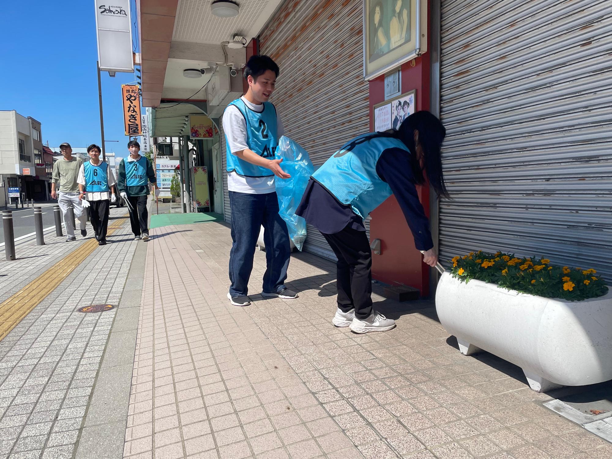 歩道に設置された花壇の後ろにあるごみをトングで拾っている女子生徒の写真