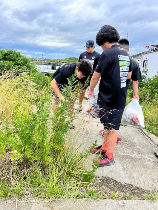蕪島の船が繋がれている海の近く。草むらのごみを探している男子学生二人の写真。