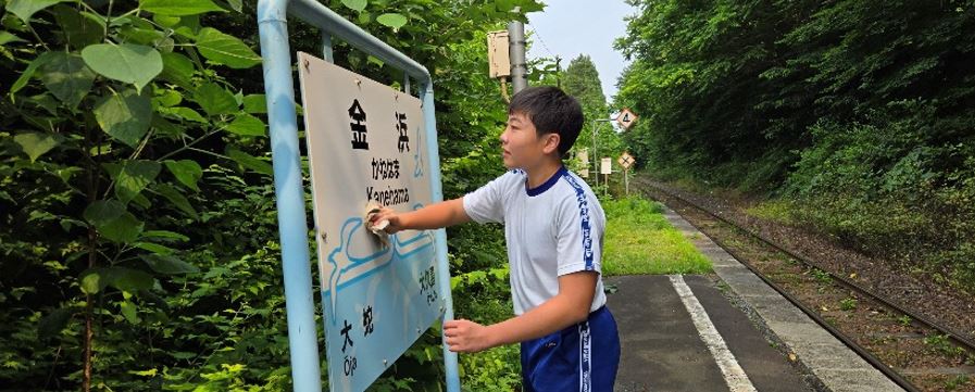 緑の多い景色の中、金浜駅の看板を拭いている男子生徒の写真