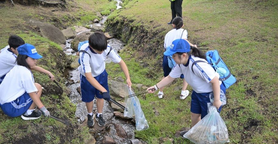 種差芝生地の小川でボランティア用ごみ袋とトングを手に持って、ごみを拾っている5人の写真