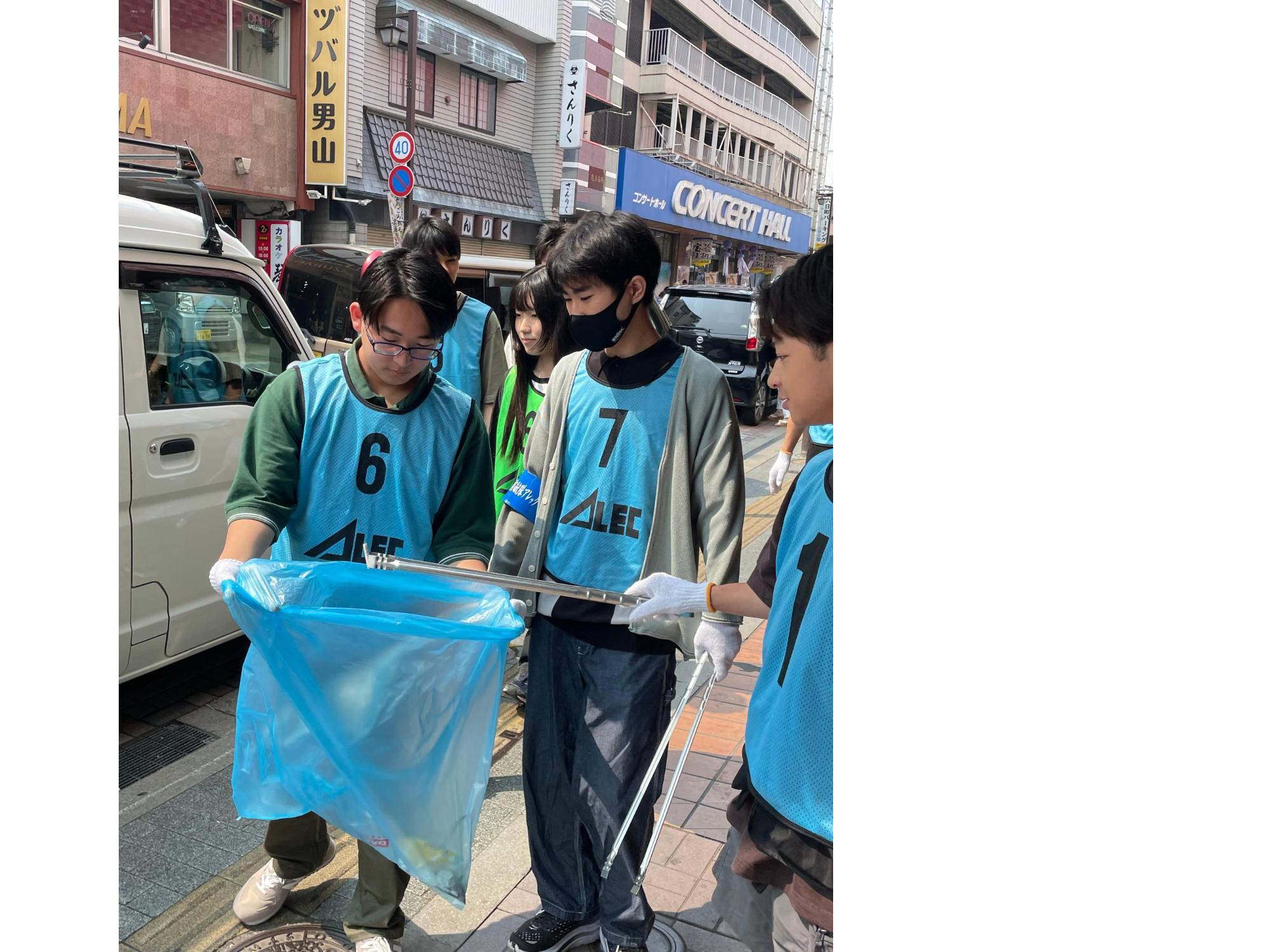 ビブスを着用し、トングでつかんだごみを別の方のひろげているごみ袋に入れている写真
