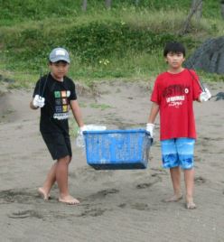 トングを持っている男児二人。青いプラスチックカゴに入ったごみを両側から持っている写真。