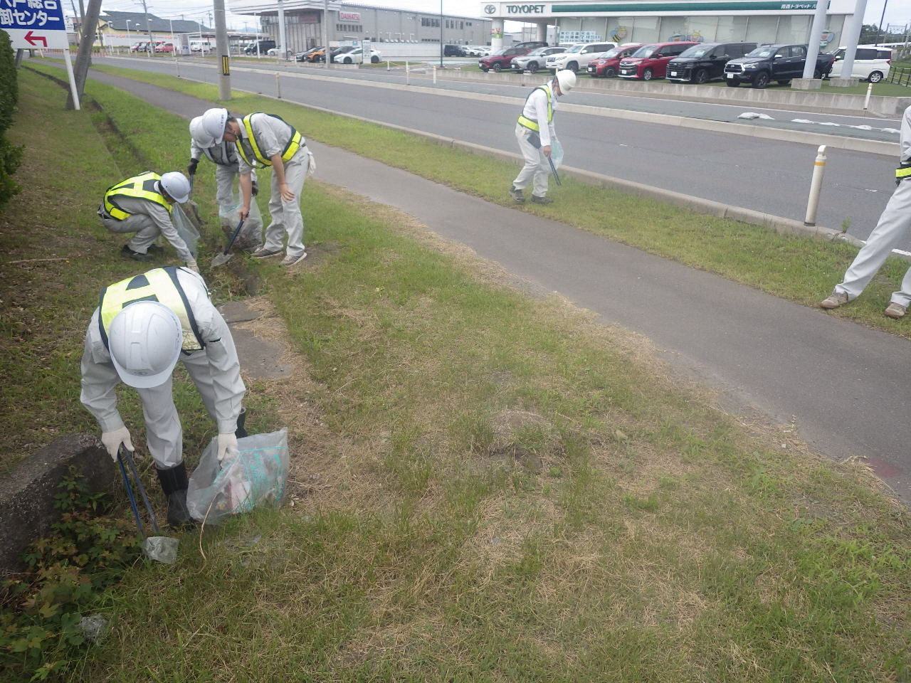 作業服にヘルメット着用で、道路脇の雑草の中のごみを拾っている写真