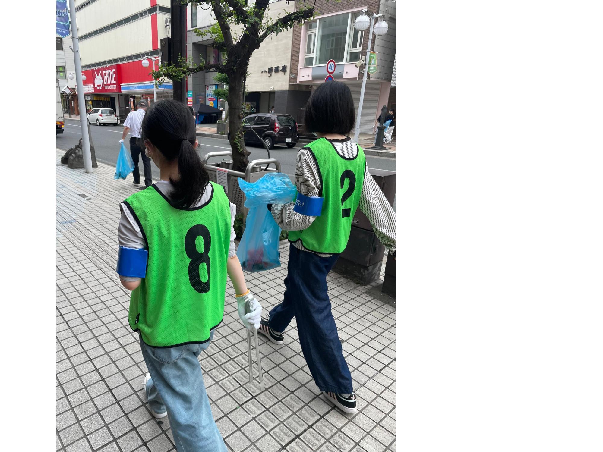 緑色のビブスを着用し、軍手にトングを持って歩道を歩く女子生徒二人の写真。