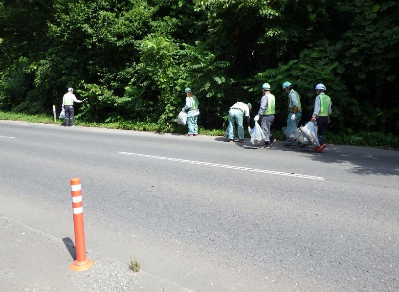 道路脇の木々の生い茂っている辺りのごみを拾っている、ヘルメット安全帯を着用した人たちの写真