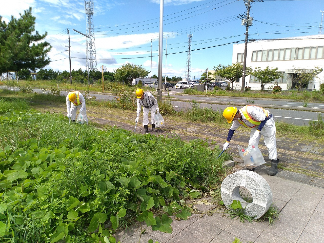 ヘルメットに作業服姿で雑草の中のごみを拾っている写真