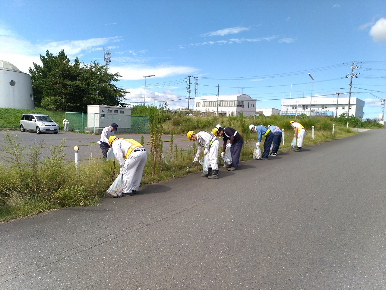 ヘルメットに作業服姿で一列に並んで、雑草のある場所のごみを拾っている写真