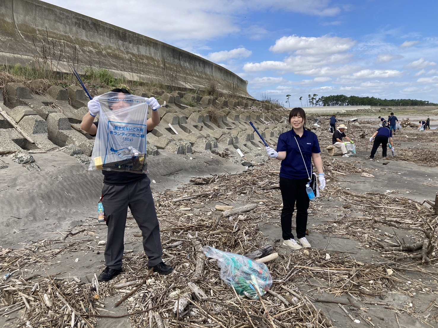 漂着した木材の広がる砂浜で、トングでごみを拾いボランティア用ごみ袋に入れている男女の写真