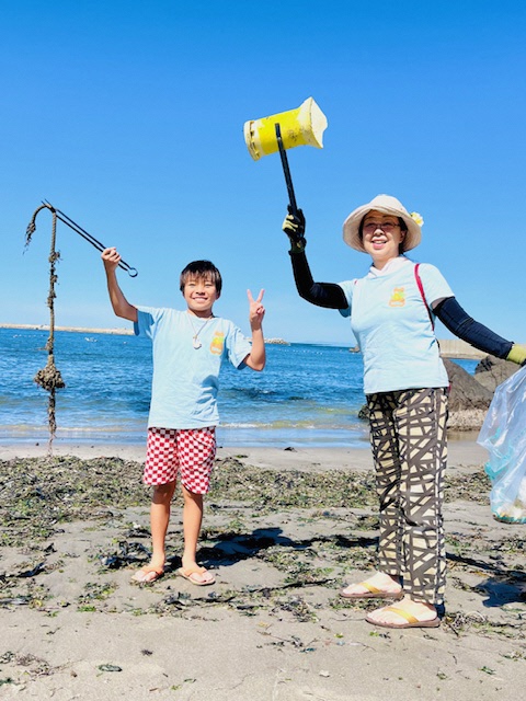 青い空と海の砂浜、トングでごみを掴んでいる男児と女性の写真