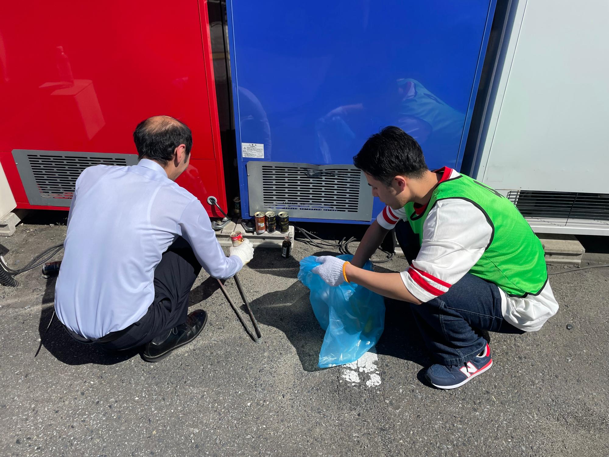 自動販売機の下のごみをしゃがんで集めている男性二人の写真