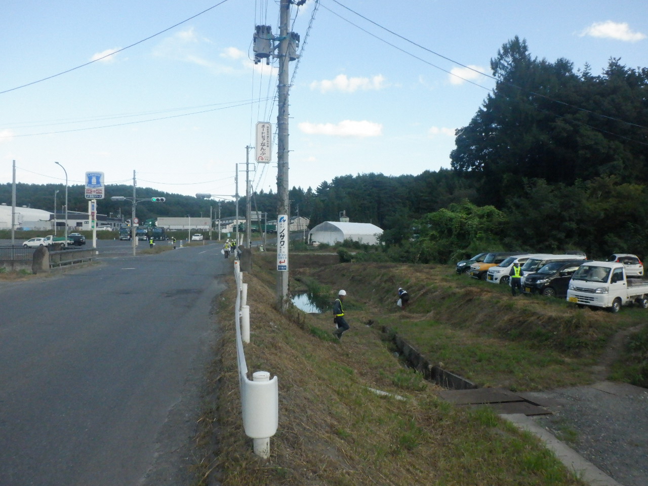 道路とガードレール下の草地の見える場所でごみ拾い活動する作業服姿の人たちの写真