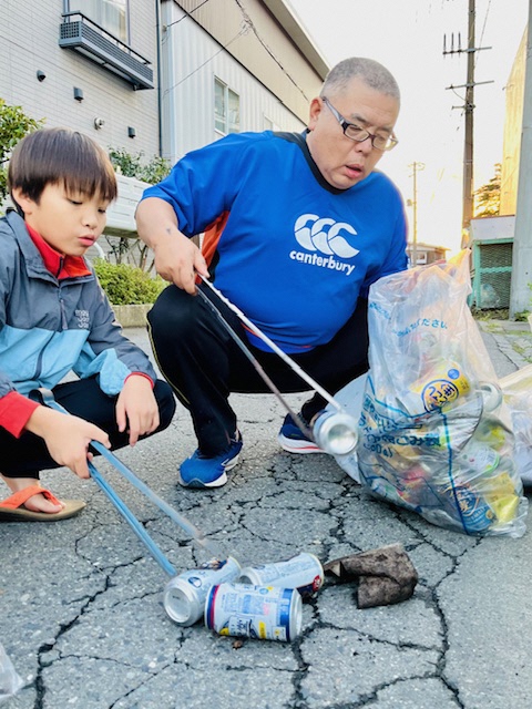 道路に落ちている飲料缶をトングで掴んでいる男性と男児の写真