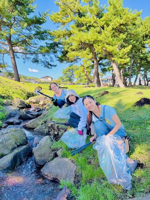 青空の広がる種差芝生地の小川。女性3人でトングでごみを集めている写真。