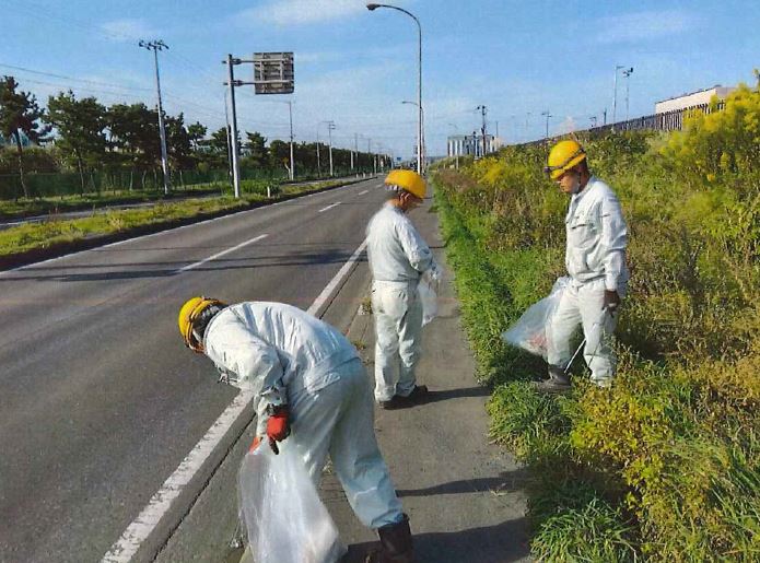 ヘルメット、作業服姿で道路脇の雑草の生えている場所のごみを集めている参加者の写真