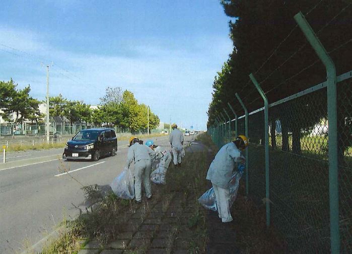 ヘルメット、作業服姿で歩道の雑草の間のごみを拾う参加者の写真