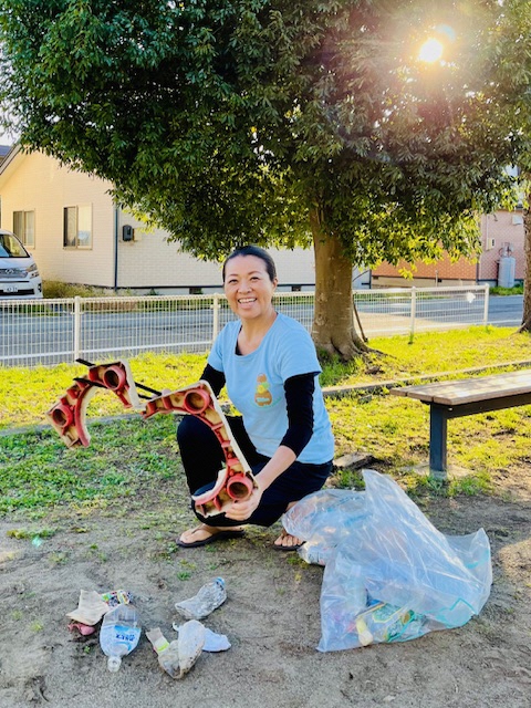 公園の中の大きな木の前。拾ったプラスチックごみをトングで掴んでいる女性の写真。