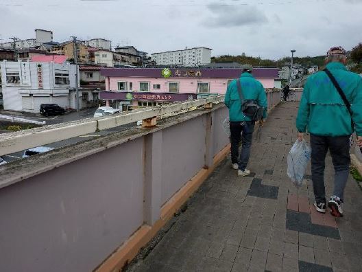 歩道橋らしき場所。同じジャンパー姿でボランティア用ごみ袋を手に持ち下を向いてごみを探している二人の写真。