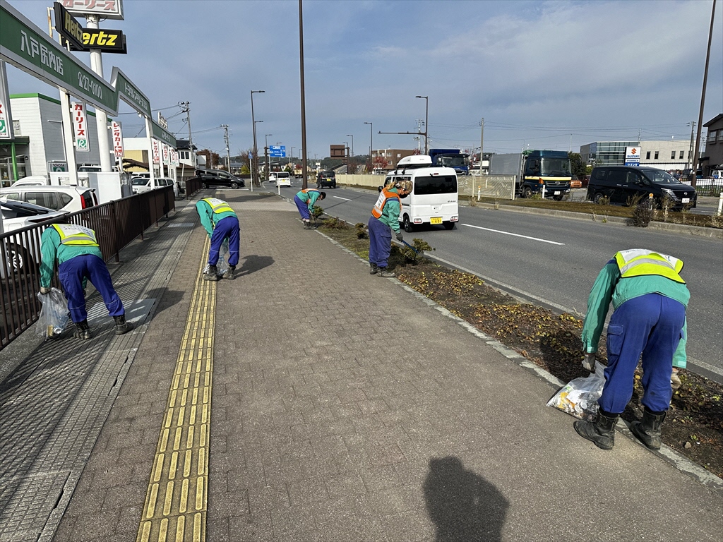 自動車販売店前の歩道で、作業服にビブスを着用し落ちているごみを拾っている5人の写真。