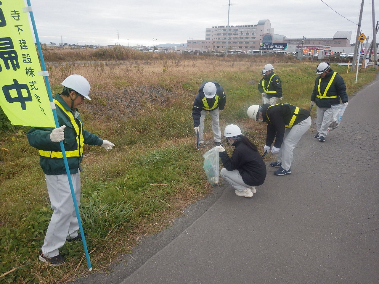 長苗代の雑草の多い歩道で、のぼり旗を掲げてヘルメット作業服姿でごみを拾う参加者の写真。