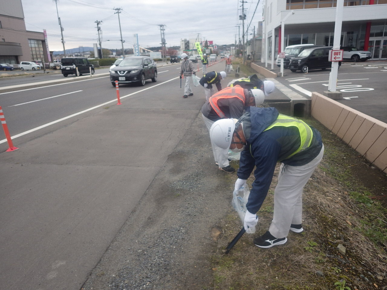 車通りの多い長苗代の道路脇。ヘルメット作業服姿で腰を曲げ、トングでごみを拾う参加者の写真。